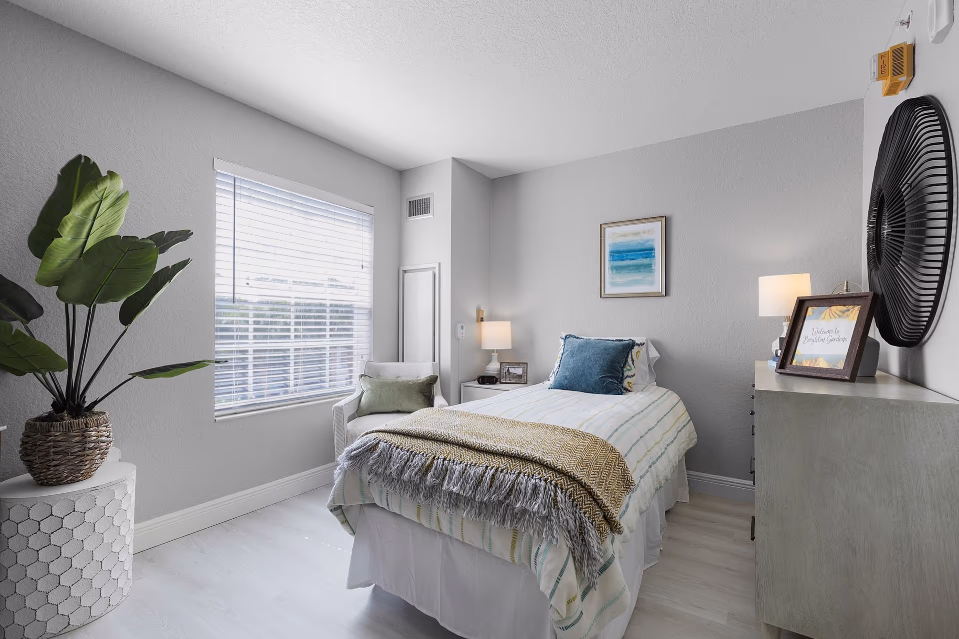 A bright and cozy bedroom with a single bed dressed in white and striped bedding, a blue pillow, and a beige throw blanket. There is a small white chair with a green cushion next to a window with white blinds. A large potted plant sits on a white hexagonal side table. Two lamps provide soft lighting, one on a nightstand beside the bed and another on a dresser. A framed abstract painting hangs above the bed, and a decorative round black wall piece is mounted above the dresser. A small framed sign on the dresser reads 'Welcome to Stratford Gardens.'