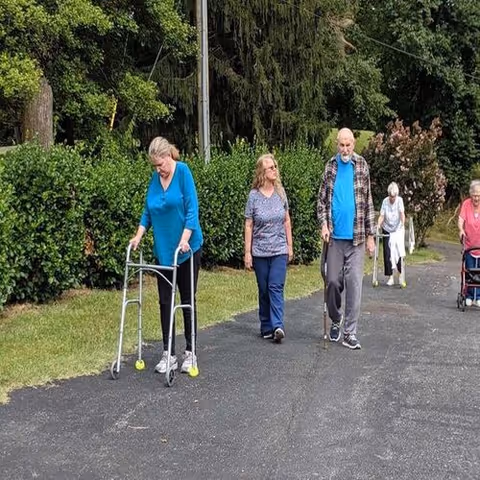 Five elderly individuals walking outdoors on a paved path surrounded by greenery. One woman in a blue top uses a walker, another woman walks beside her, an elderly man with a cane walks next to them, and two more elderly women walk behind them, one using a walker and the other walking unaided.