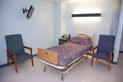A hospital-style room with a single bed covered in a purple patterned blanket, flanked by two wooden chairs with blue cushions. A wall-mounted TV is positioned above one chair, and a small bedside table with medical supplies is next to the bed. The room has light-colored walls and flooring.
