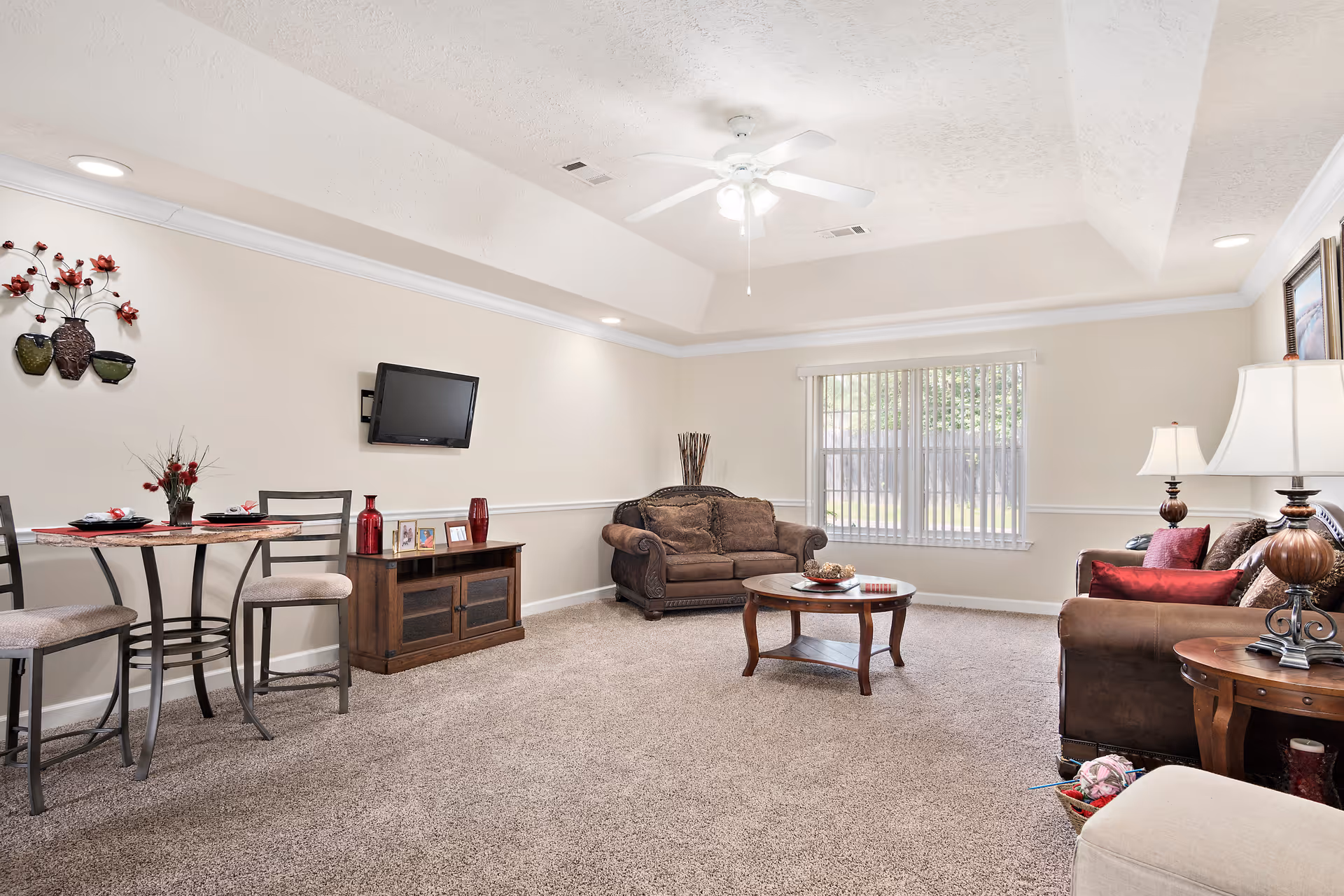 A bright living room with beige walls and carpeted floor. The room features two brown upholstered sofas with decorative pillows, a wooden coffee table with a bowl of decorative items, a small wooden cabinet with framed photos and red vases, a wall-mounted flat-screen TV, a round dining table with four chairs, and a ceiling fan with lights. A large window with vertical blinds allows natural light into the room.