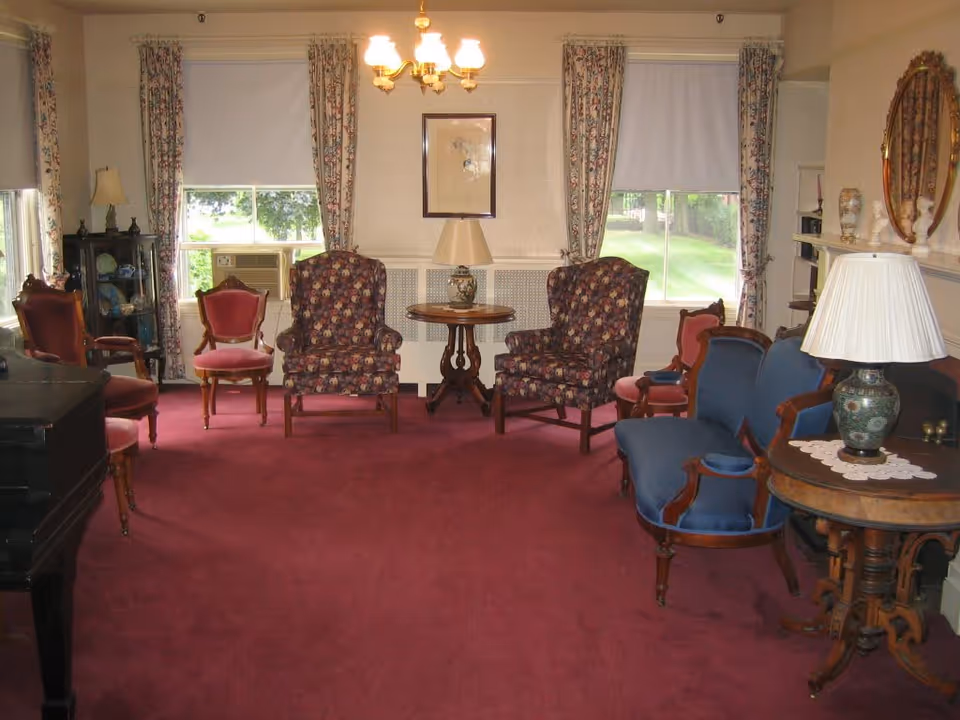A traditional living room with a burgundy carpet, floral patterned armchairs, a blue upholstered loveseat, wooden side tables with lamps, floral curtains on windows, and a chandelier hanging from the ceiling.