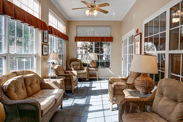 Sunlit sitting area with wicker furniture including cushioned chairs and a loveseat, several table lamps, large windows with red valances, a ceiling fan with lights, and a tiled floor.