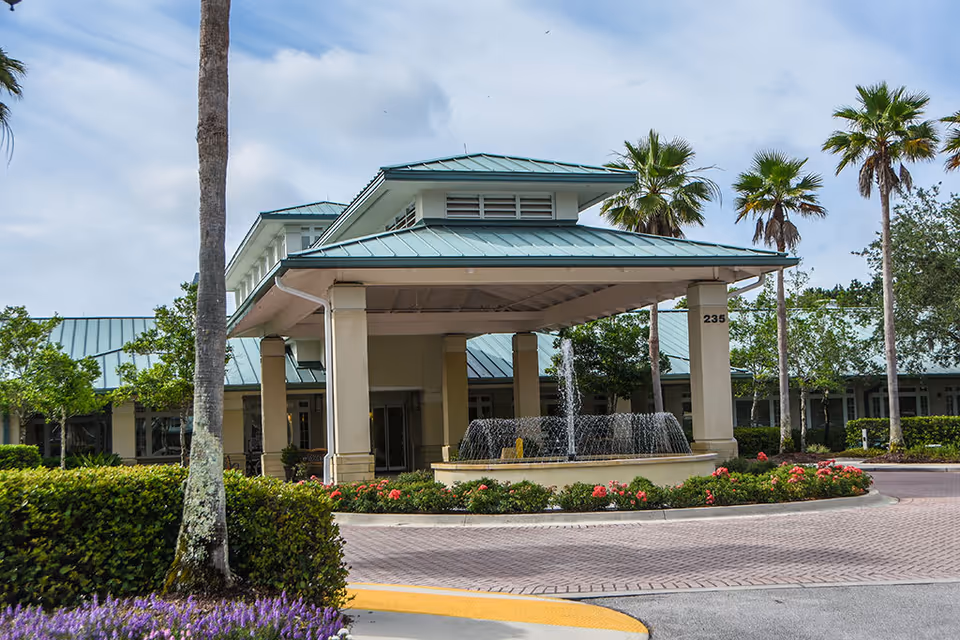 Entrance of Westminster St. Augustine facility with a covered driveway, a water fountain in front, surrounded by palm trees, bushes, and flowers under a partly cloudy sky.