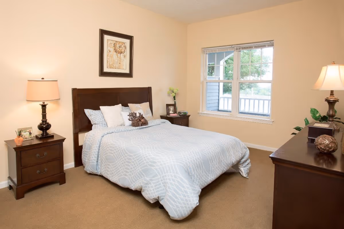 A neatly made bed with light blue and white patterned bedding in a bedroom. The room has beige walls and carpet, two wooden nightstands with lamps on either side of the bed, a framed floral picture above the headboard, a window with a view of greenery outside, and a wooden dresser with a lamp and decorative items.