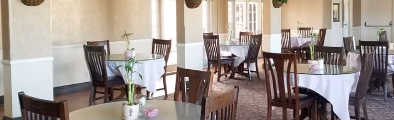 Bright dining room with round tables draped in white tablecloths, wooden chairs, and small potted plant centerpieces.