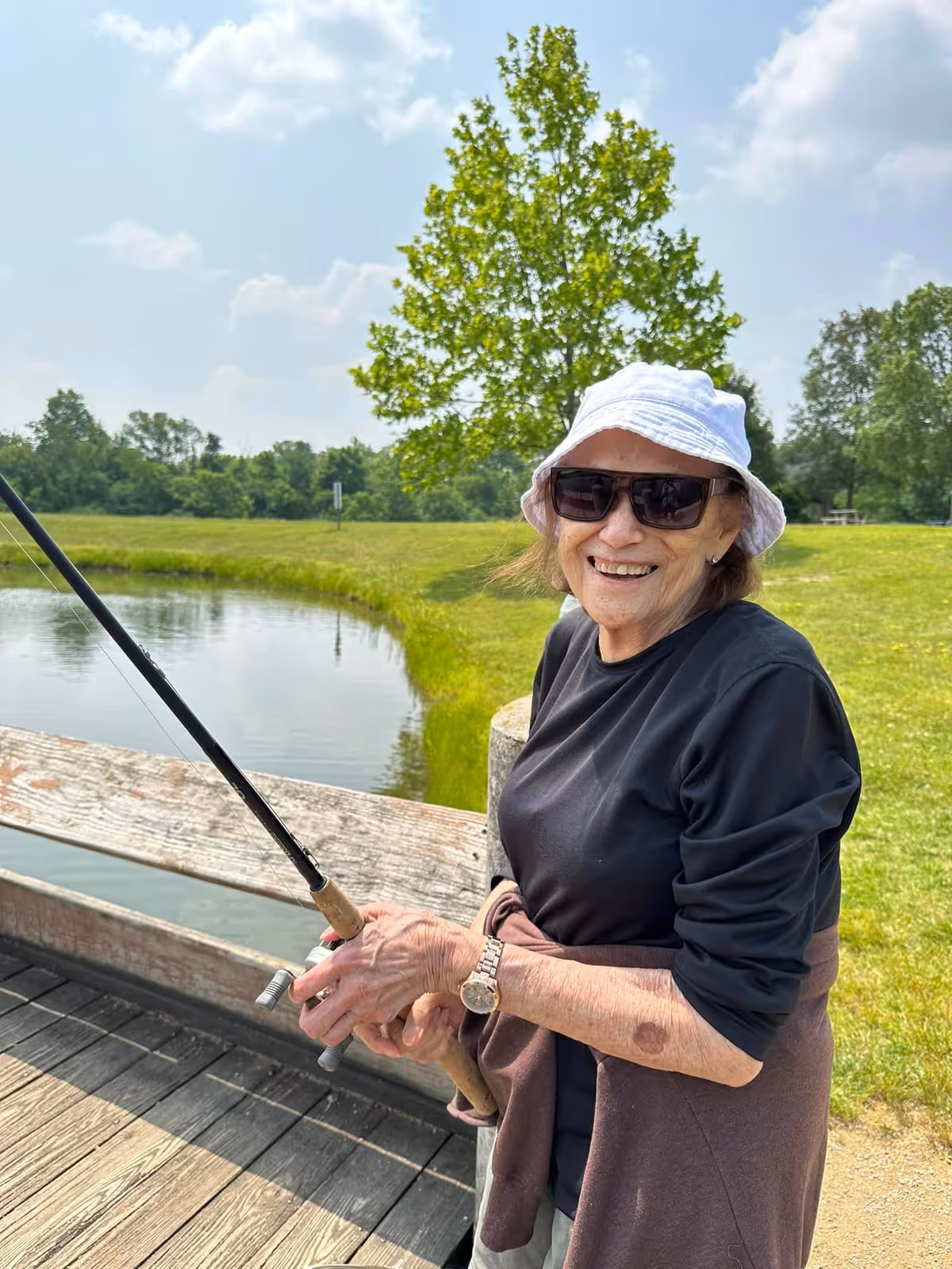 An elderly woman wearing a white sun hat, dark sunglasses, and a black long-sleeve shirt is smiling while holding a fishing rod on a wooden dock by a pond. The background features green grass, trees, and a partly cloudy sky.