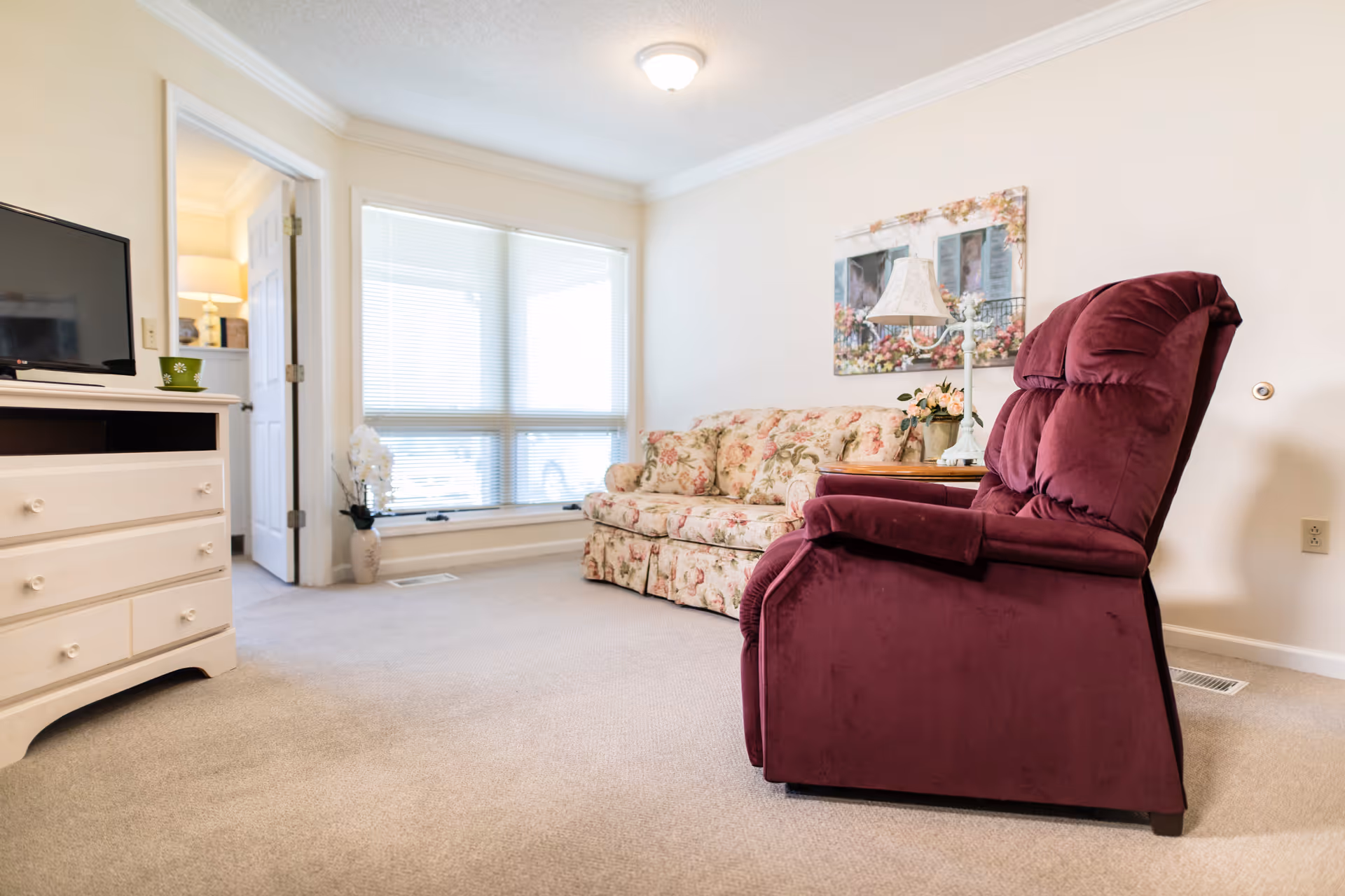 A cozy living room with a floral patterned sofa, a burgundy recliner chair, a white dresser with a TV on top, a side table with a lamp and flowers, and large windows with blinds letting in natural light.