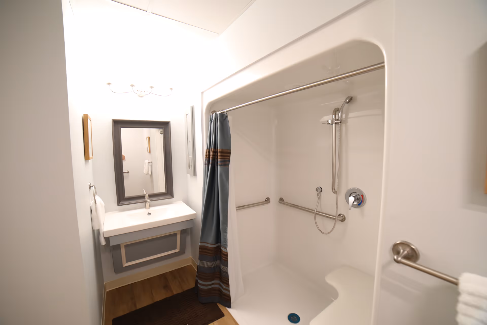 A clean, accessible bathroom featuring a walk-in shower with grab bars and a handheld showerhead, a shower curtain with blue and brown stripes, a white sink with a modern faucet, a rectangular mirror with a dark frame, and a towel hanging on a ring next to the sink. The floor has wood-like flooring and a dark brown bath mat.