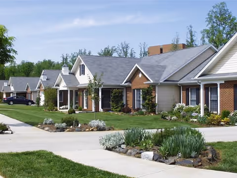Row of single-story residential buildings with brick and siding exteriors, surrounded by well-maintained lawns, shrubs, and small trees under a clear blue sky.