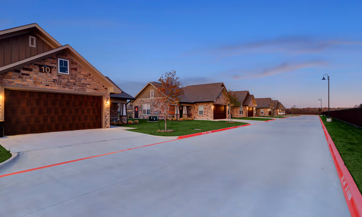 A row of single-story residential buildings with stone facades and wooden garage doors along a wide concrete driveway at dusk, with street lamps and small trees planted in grassy areas.