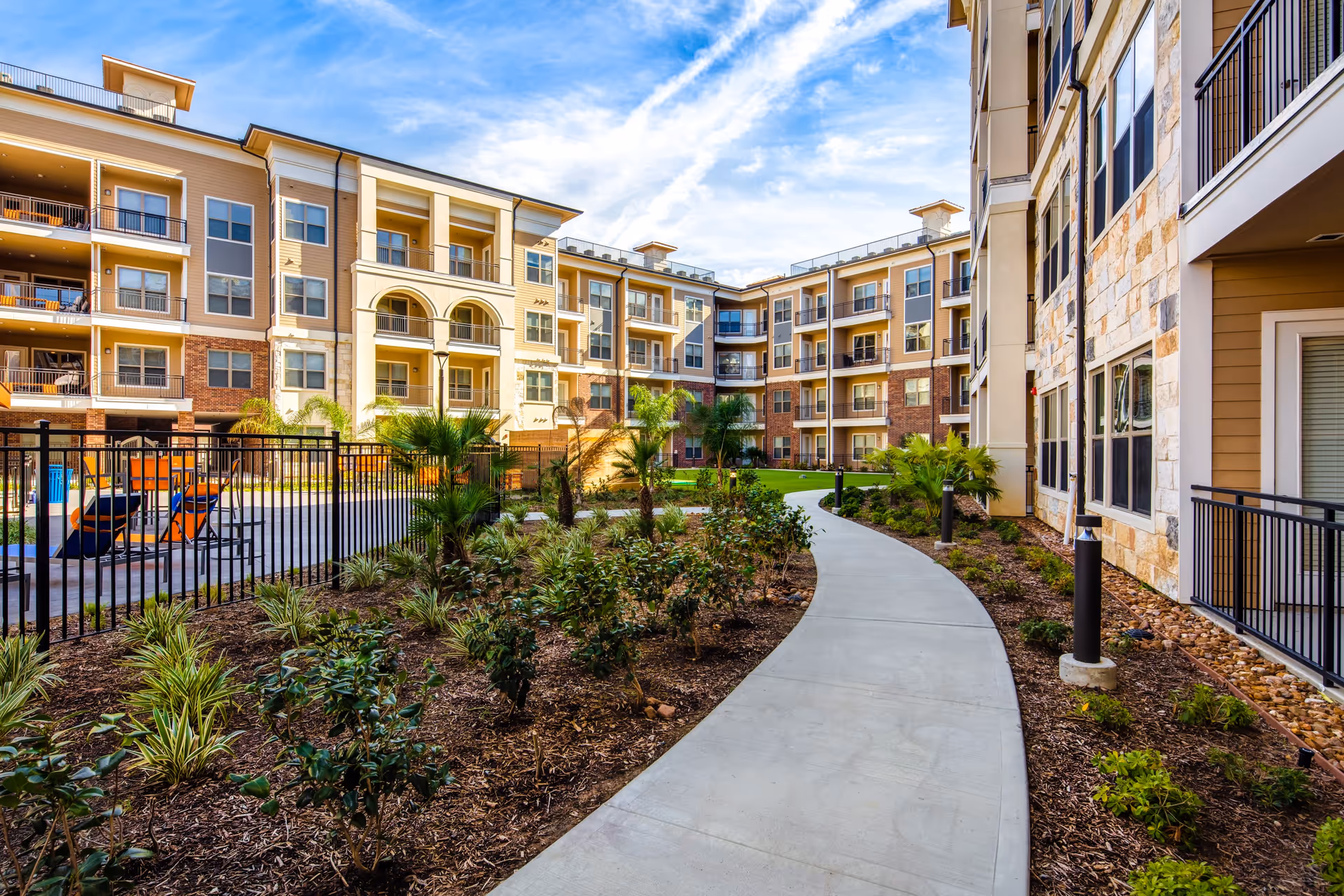 Landscaped courtyard with a curved walkway between multi-story senior living buildings, balconies, and a fenced pool area.