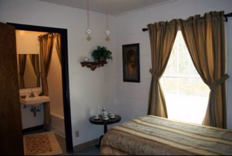 A small bedroom with a bed covered in a patterned bedspread, a window with brown curtains tied back, a small round table with a tea set, a wall shelf with a plant and decorative items, and an open door leading to a bathroom with a sink, mirror, and shower curtain.