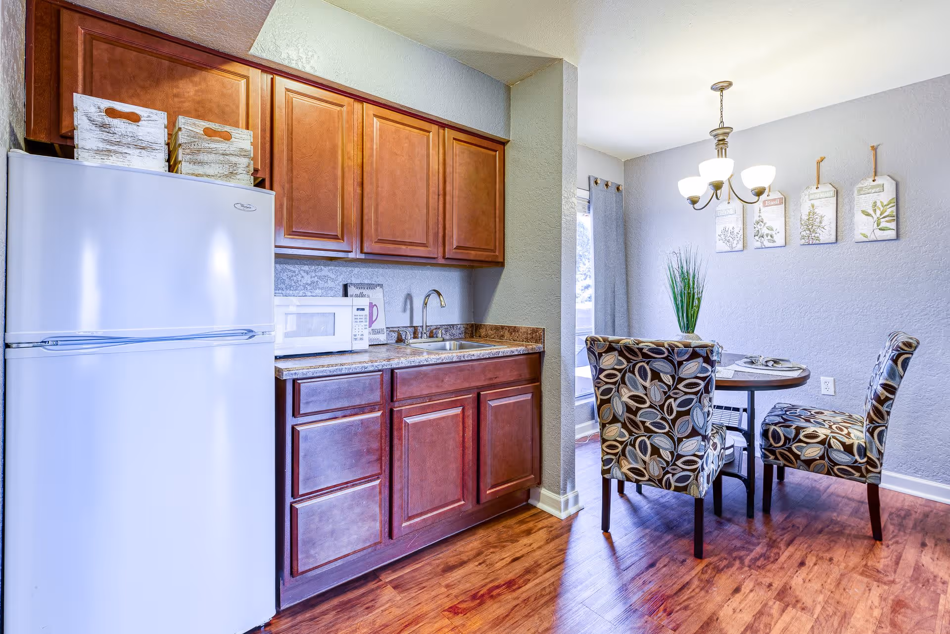 A small kitchen area with wooden cabinets, a white refrigerator, a microwave on the countertop, and a sink. Adjacent to the kitchen is a dining area with a round table and four patterned upholstered chairs. A chandelier hangs above the table, and decorative wall art is displayed on the gray wall. The floor is wooden.
