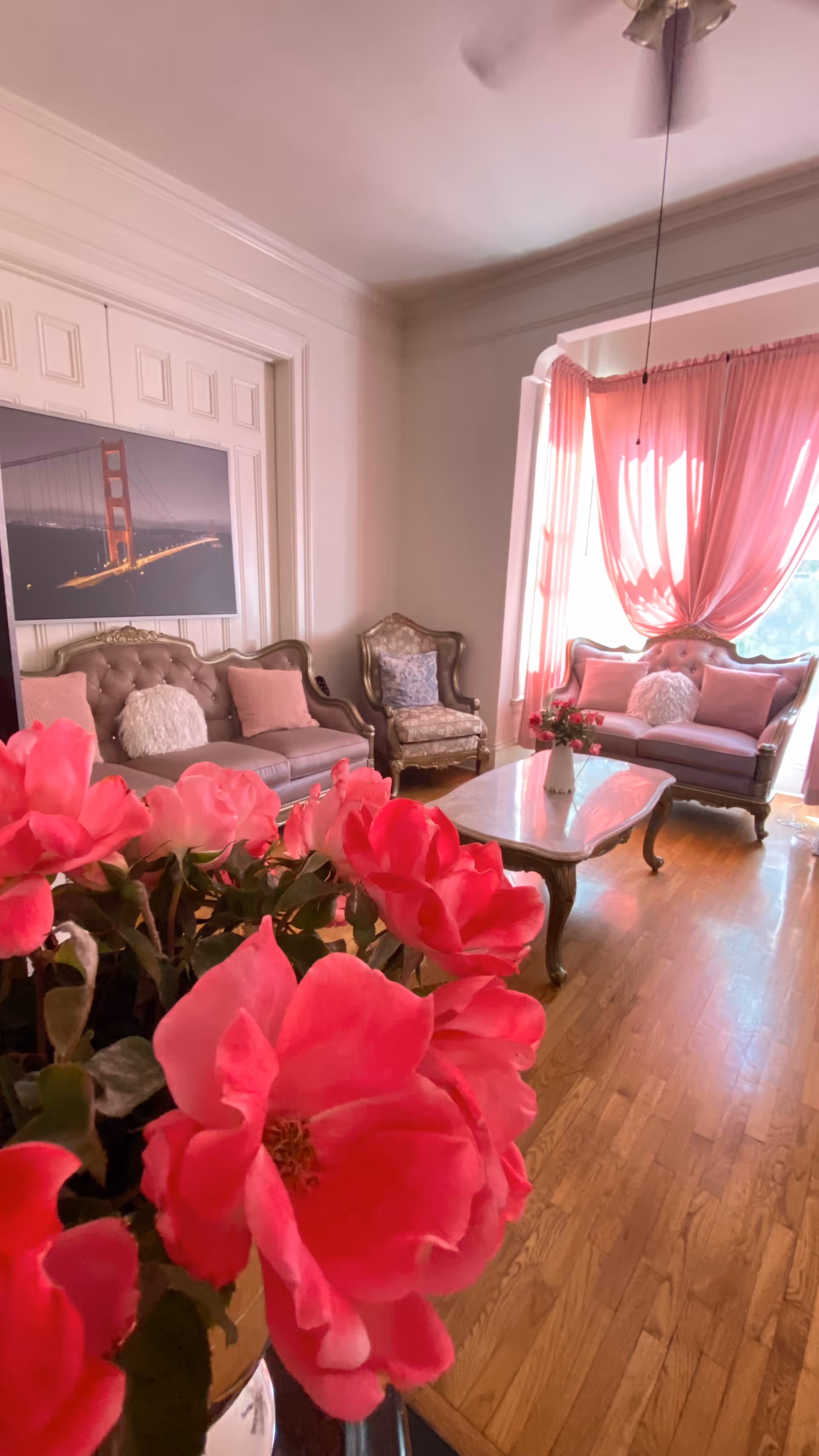 A cozy living room with vintage-style furniture including two tufted sofas and an armchair, all adorned with decorative pillows. A white coffee table sits in the center on a wooden floor. Pink curtains drape over a large window, allowing natural light to fill the room. In the foreground, a vase of vibrant pink flowers is prominently displayed. A framed picture of the Golden Gate Bridge hangs on the wall.