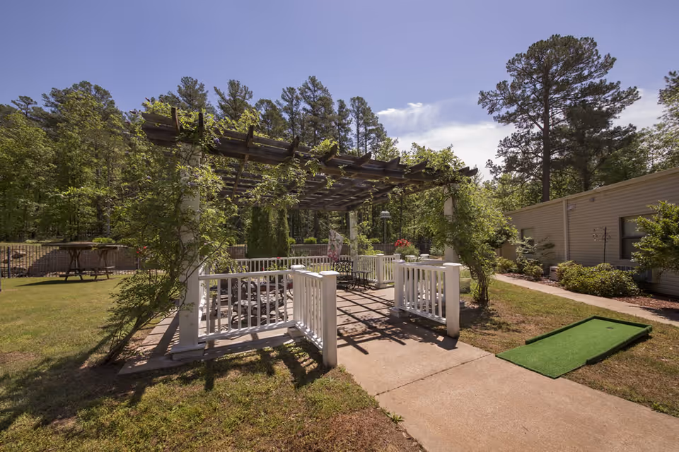 Outdoor garden area at Perry County Nursing & Rehab Center featuring a pergola with climbing plants, white railings, patio furniture underneath, a putting green on the right, and surrounded by trees and greenery under a clear blue sky.