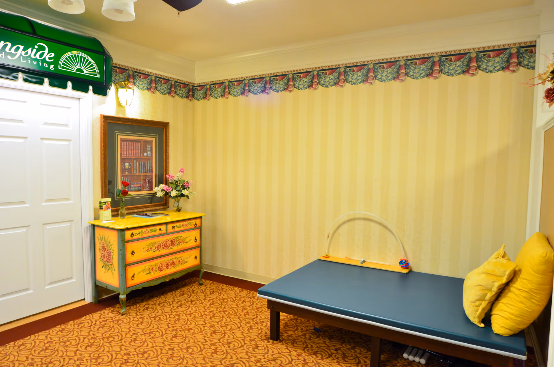 A cozy room with yellow striped wallpaper and a patterned carpet. There is a blue padded bench with two yellow pillows on the right side. On the left side, there is a decorative yellow chest of drawers with floral designs, topped with a vase of flowers and a framed picture of books on the wall above it. A green awning with partial text 'ngside Living' is visible above white double doors on the left.