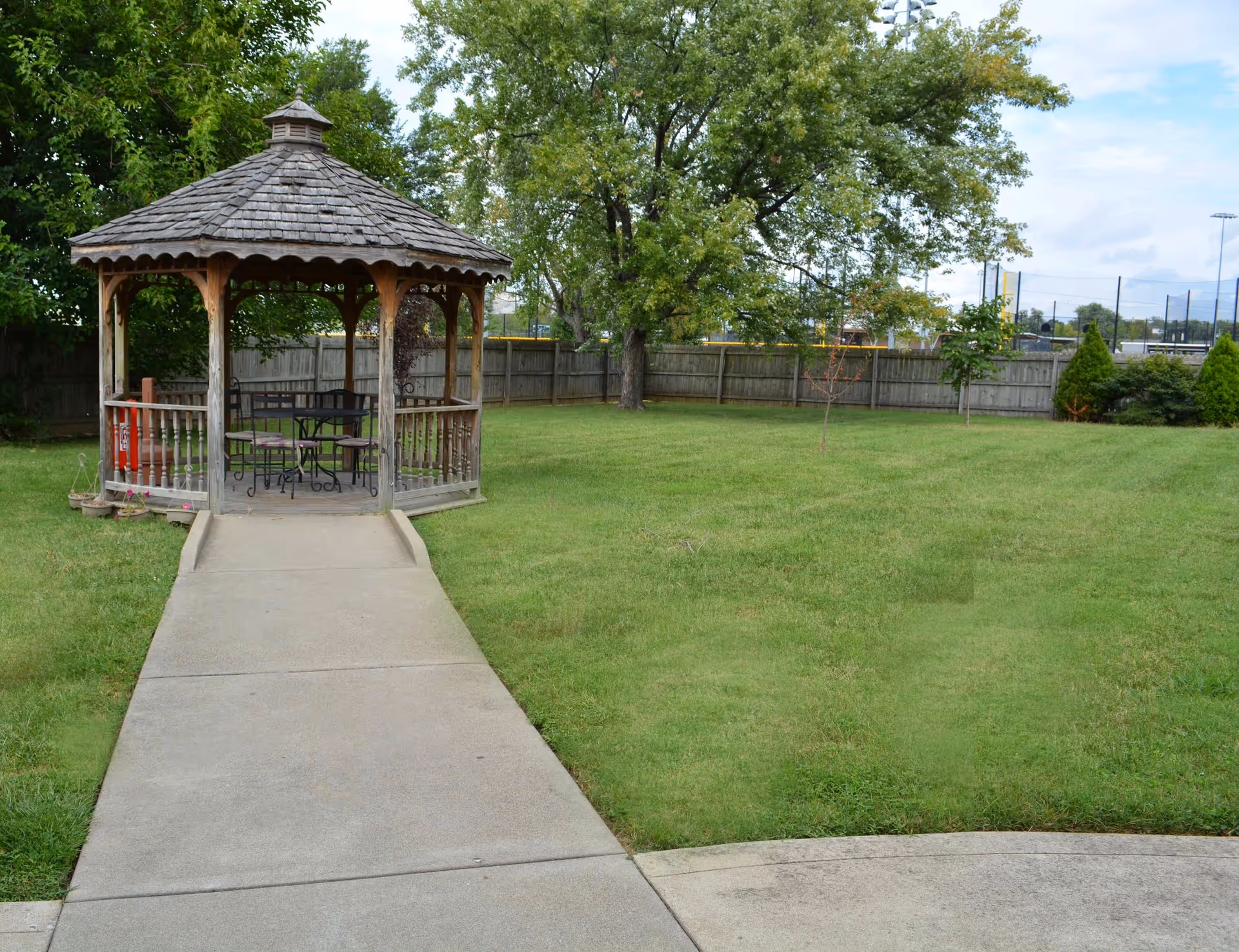 A wooden gazebo with a shingled roof sits at the end of a concrete pathway in a grassy yard. Inside the gazebo are a metal table and chairs. The yard is enclosed by a wooden fence and has several trees and shrubs.