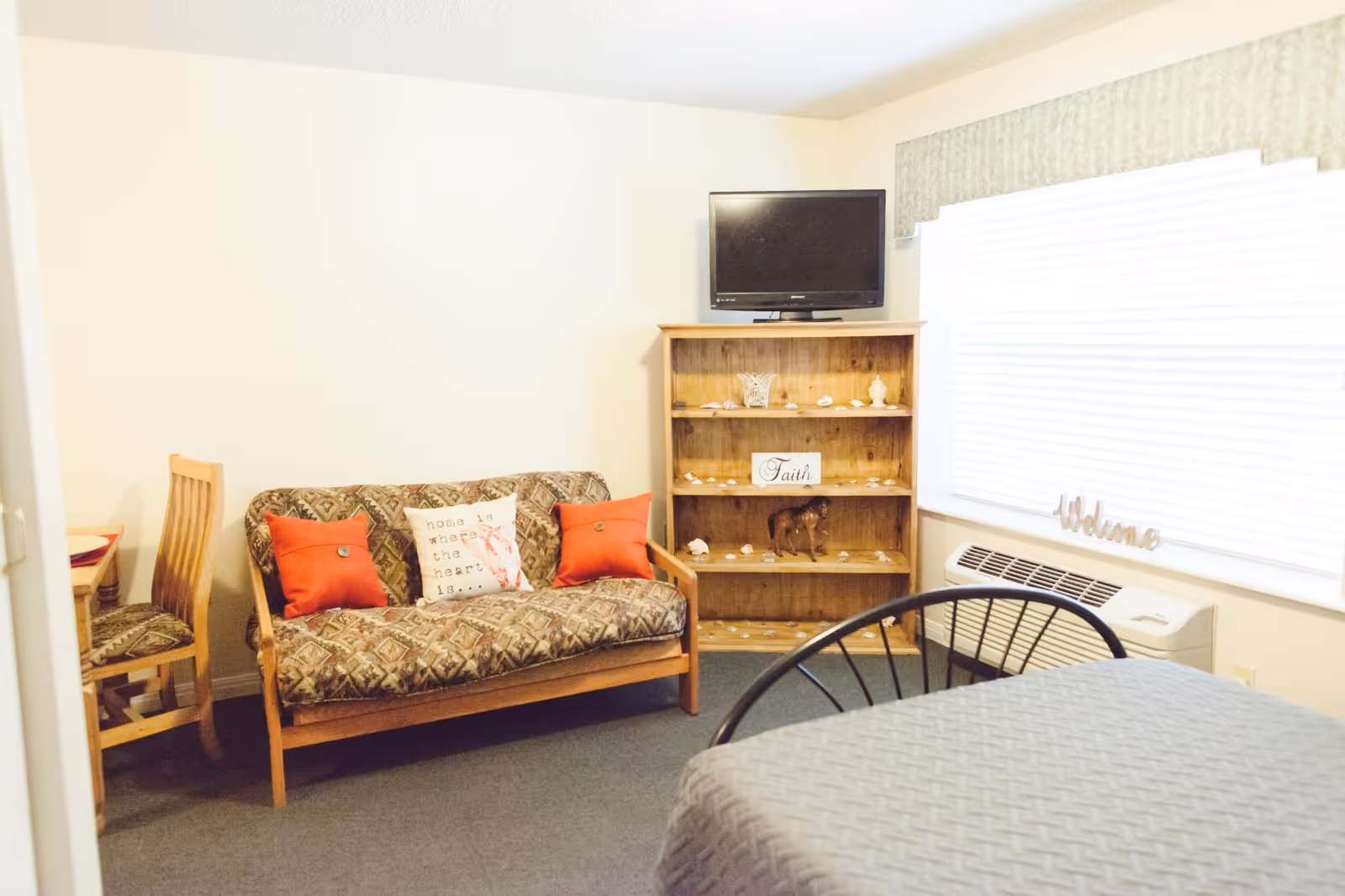 Cozy sitting area with a patterned loveseat and red pillows, a wooden bookshelf topped by a TV, and a window with blinds and an air conditioner.