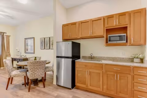 A small kitchen area with wooden cabinets, a granite countertop, a stainless steel refrigerator, and a microwave. Adjacent to the kitchen is a dining area with a round table and four patterned upholstered chairs. The room has light-colored walls and wood flooring, with framed artwork on the wall and a window with curtains in the background.