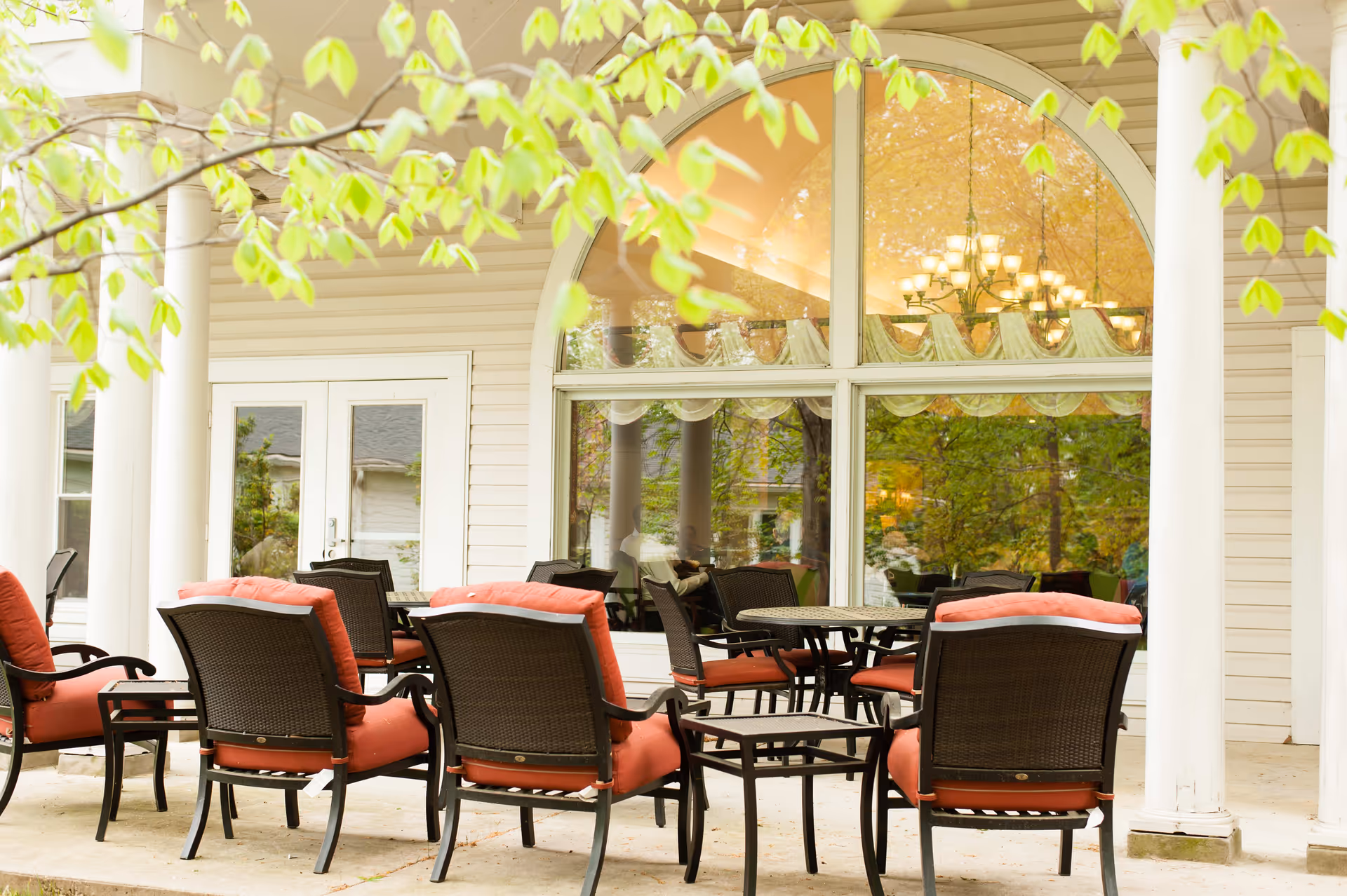 Outdoor patio area with several black wicker chairs with red cushions arranged around small tables. The patio is adjacent to a building with large arched windows and white pillars, with green leaves from nearby trees partially visible in the foreground.