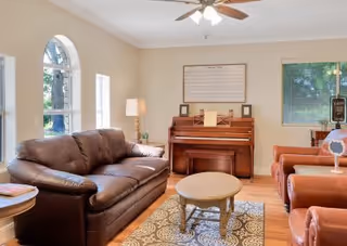Well-lit living room with leather sofas and armchairs, a piano against the wall, a round coffee table, rug, and ceiling fan.