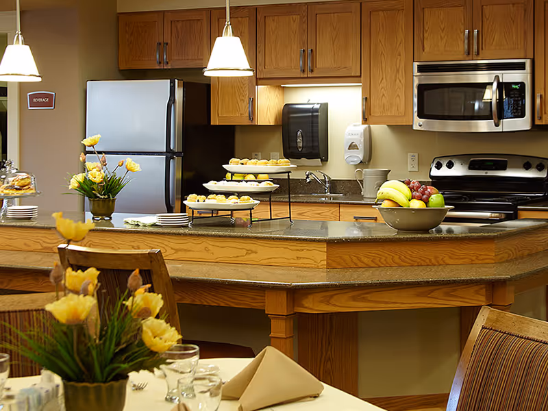 A kitchen area in a senior living facility with wooden cabinets, a stainless steel refrigerator, microwave, and stove. The countertop has a tiered tray with pastries and a bowl of fruit. There are yellow flowers in vases on the counter and dining table, with neatly folded napkins and glassware set on the table.
