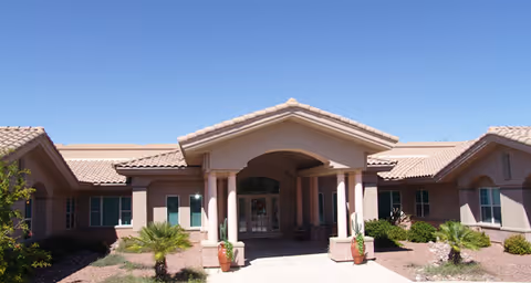 Front exterior view of a single-story senior living facility building with a tiled roof, a covered entrance supported by columns, desert landscaping including small palm trees and cacti, under a clear blue sky.