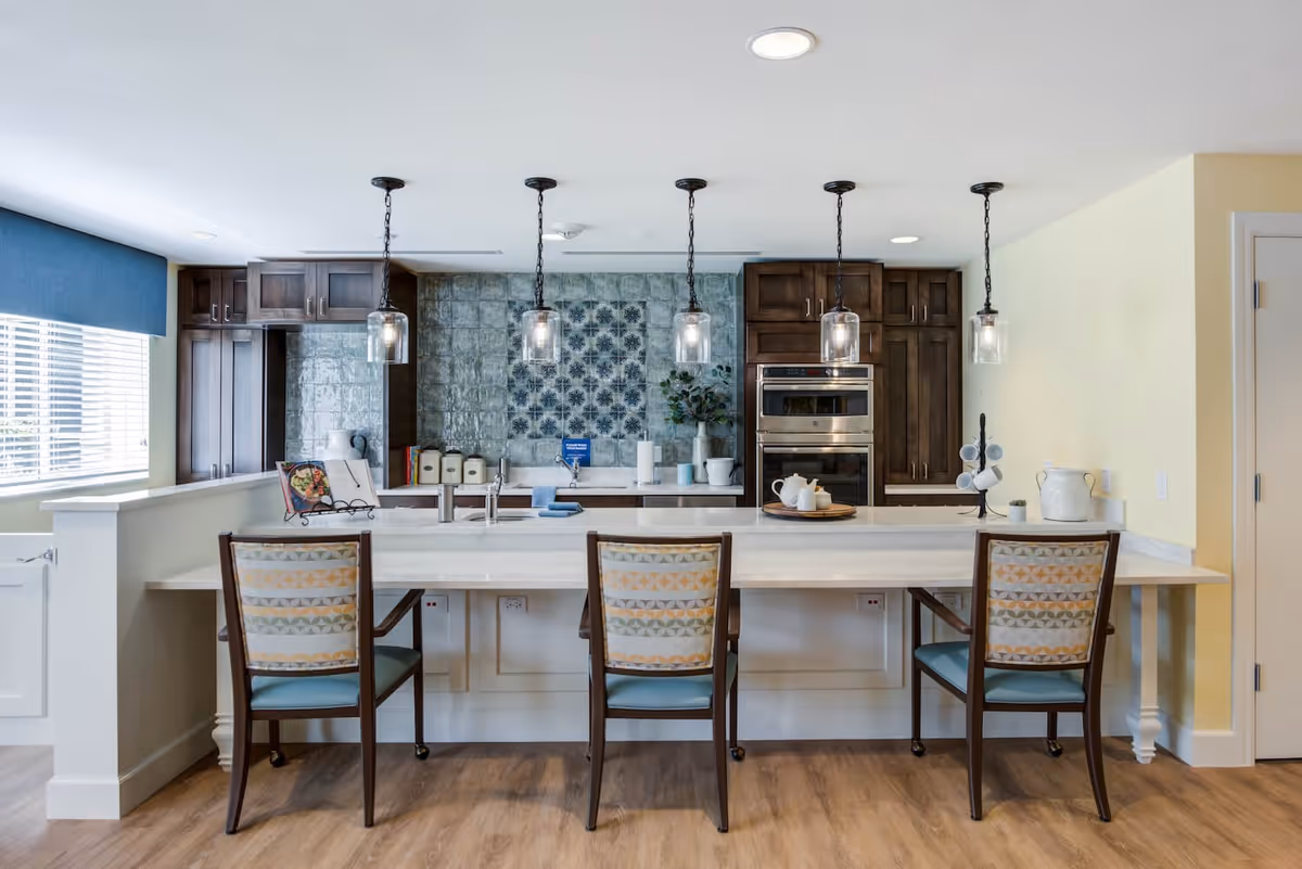 A modern kitchen area with a long white countertop island featuring three chairs with patterned backs and blue seats. Above the island hang five pendant lights with glass shades. The kitchen has dark wooden cabinets, a built-in oven, a tiled backsplash with a decorative blue pattern, and various kitchen items including a cookbook, canisters, and a tea set. The floor is wood, and there is a window with blinds and a blue valance on the left side.