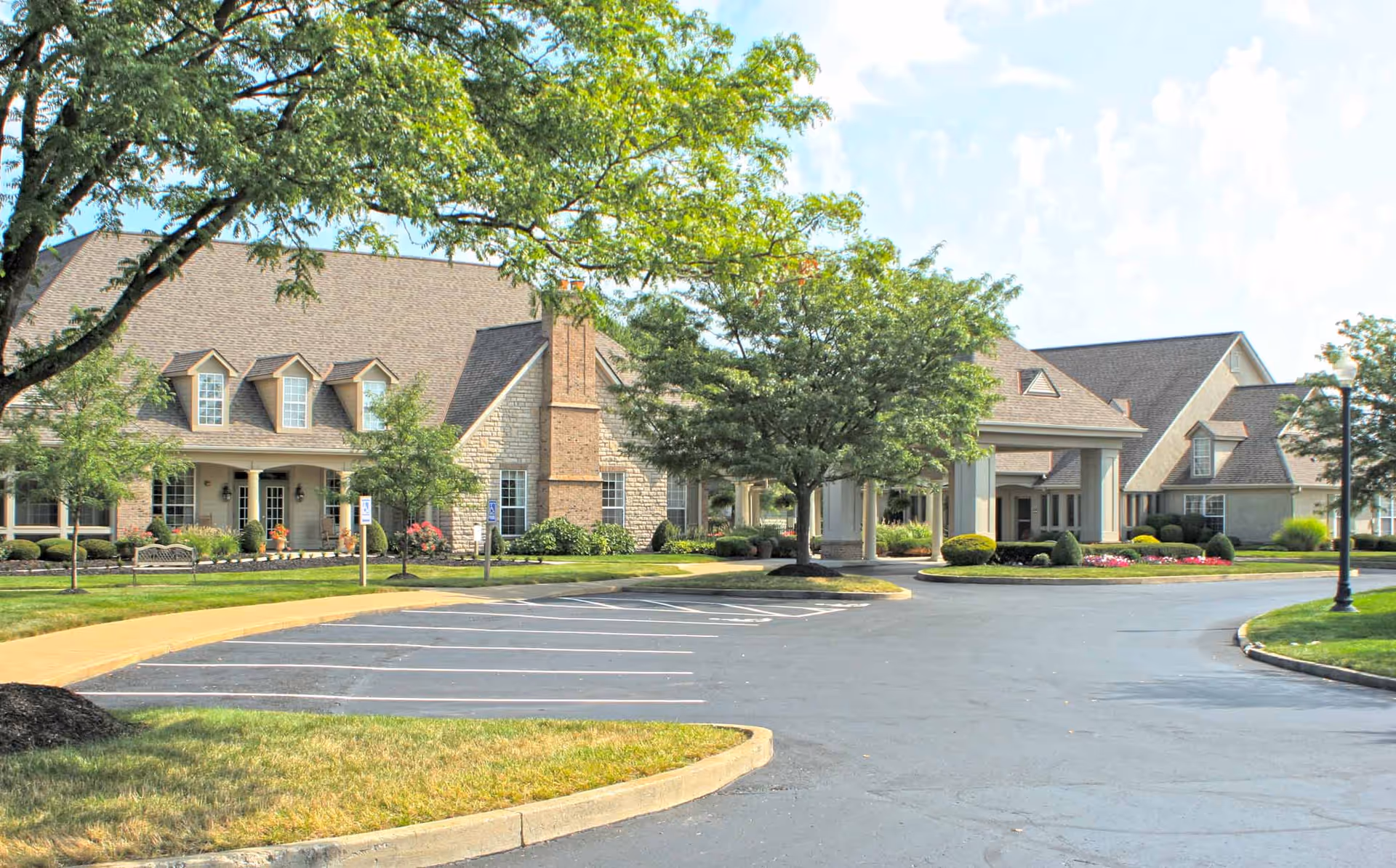 Exterior view of Feridean Commons senior living facility showing a large building with a pitched roof, dormer windows, and a covered entrance. The foreground features a paved parking lot with marked spaces, landscaped greenery, trees, and a clear sky.