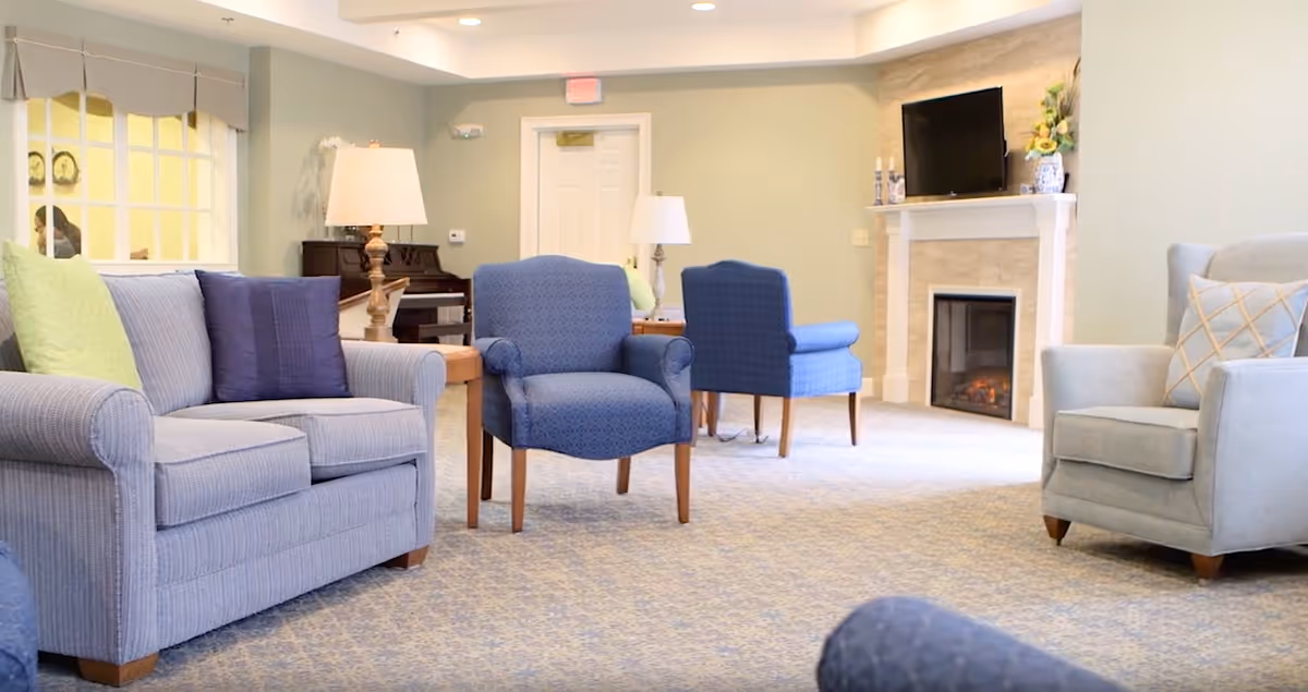 A cozy living room area with a light blue loveseat, two blue armchairs, and a gray armchair with a decorative pillow. There are two wooden side tables with lamps, a fireplace with a TV mounted above it, and a piano in the background. The room has light-colored walls and carpeted flooring.