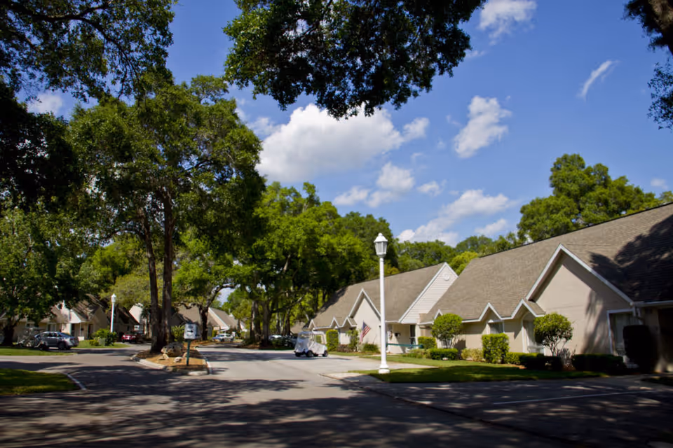 A sunny street view of a residential area in John Knox Village of Central Florida with single-story houses, green trees, and a clear blue sky with some clouds.