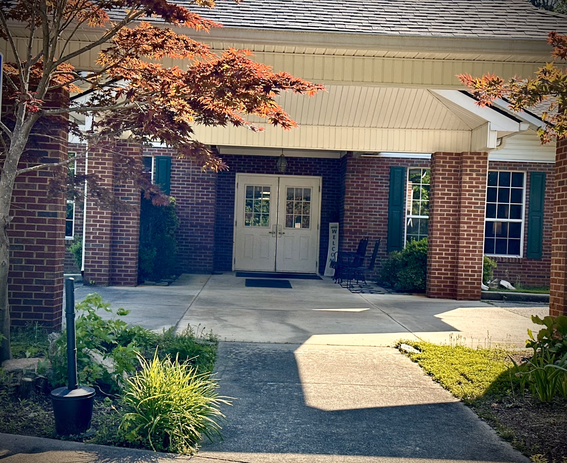 Entrance to a brick building with double white doors under a covered porch. There are green plants and shrubs along the walkway leading to the entrance, and a tree with reddish leaves partially shading the area. Two chairs and a welcome sign are visible near the door.