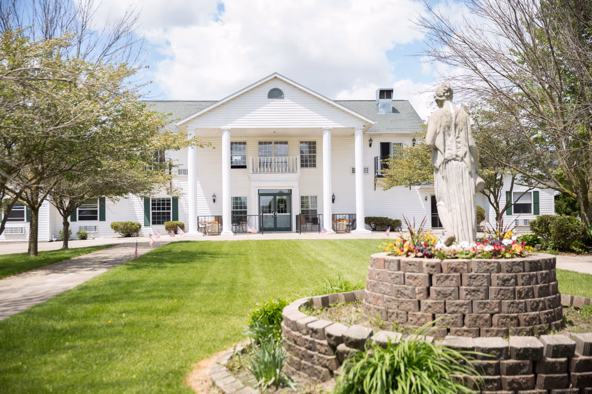 Front exterior view of a white two-story building with large white columns and a small balcony above the entrance. The building is surrounded by green grass, trees, and a circular brick flower bed with a statue in the foreground. The sky is partly cloudy.