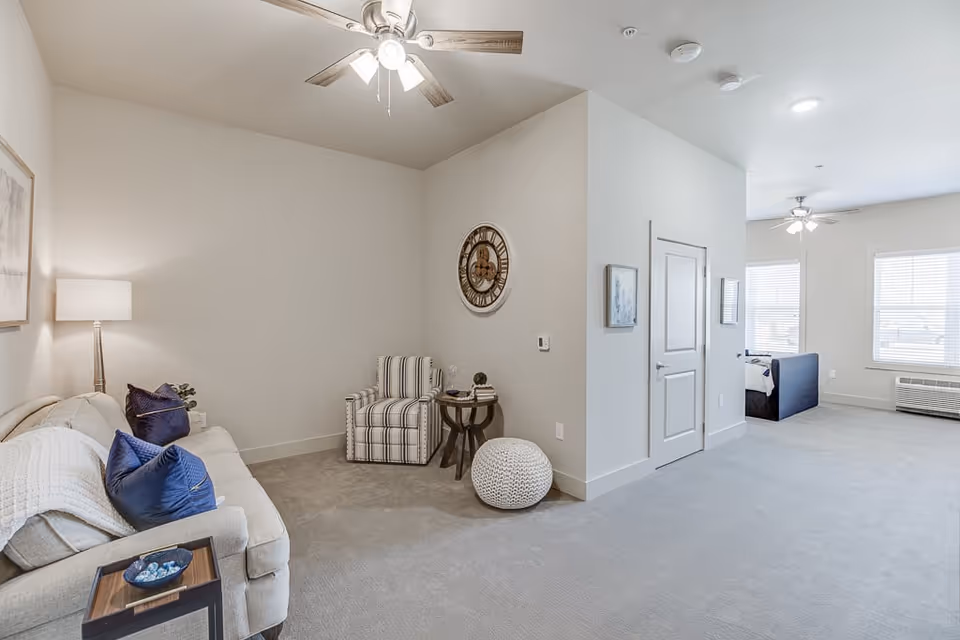 A bright and spacious living area in an assisted living facility featuring a beige sofa with blue pillows, a striped armchair, a round side table, a white knitted pouf, a floor lamp, and a large wall clock. The room has light-colored walls and carpet, ceiling fans with lights, and large windows allowing natural light to enter.
