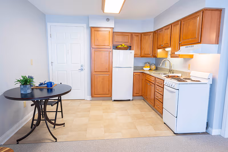 A small kitchen area in a senior living facility with wooden cabinets, a white refrigerator, a white stove with an overhead vent, and a countertop with a sink. To the left, there is a round black table with a plant, a blue mug, and some papers on it. The floor is tiled, and the walls are painted light blue. A white door is visible in the background.