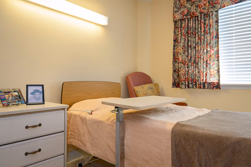 A small bedroom in a senior living facility with a single bed covered in a light pink bedspread and a brown blanket at the foot. There is a bedside table with two framed photos on it. A red armchair with a floral cushion is positioned in the corner near a window with floral curtains and white blinds. A wall-mounted light fixture illuminates the room.