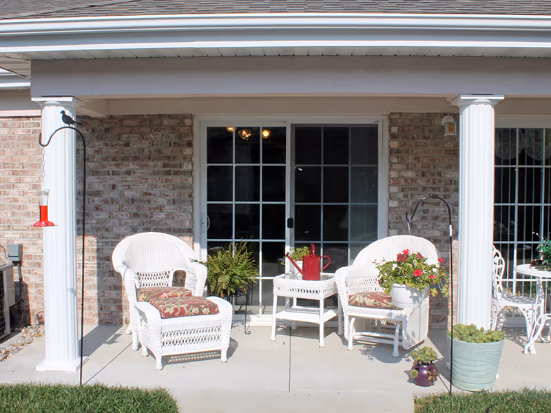 Covered patio with white wicker chairs, a small table, potted plants, and sliding glass doors set in a brick wall.