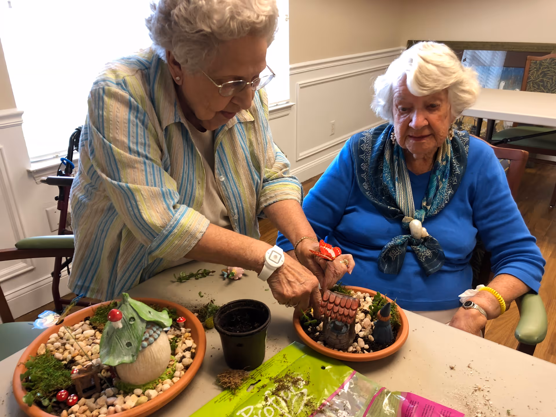 Two elderly women sitting at a table engaged in a miniature gardening activity. One woman is wearing a striped shirt and glasses, arranging items in a small pot, while the other woman in a blue top and scarf watches. The table has two small pots with decorative miniature garden scenes, a small black pot, and a bag of potting soil.