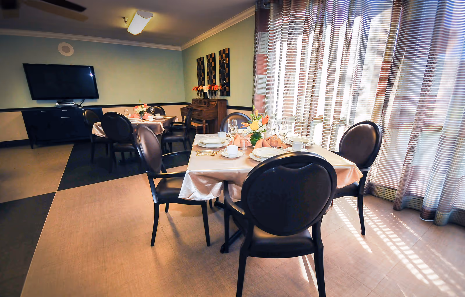 A dining room with tables set for a meal, including plates, cups, glasses, and floral centerpieces. The room has large windows with sheer curtains allowing natural light to enter. There is a television mounted on the wall and a piano against the far wall with decorative artwork above it.