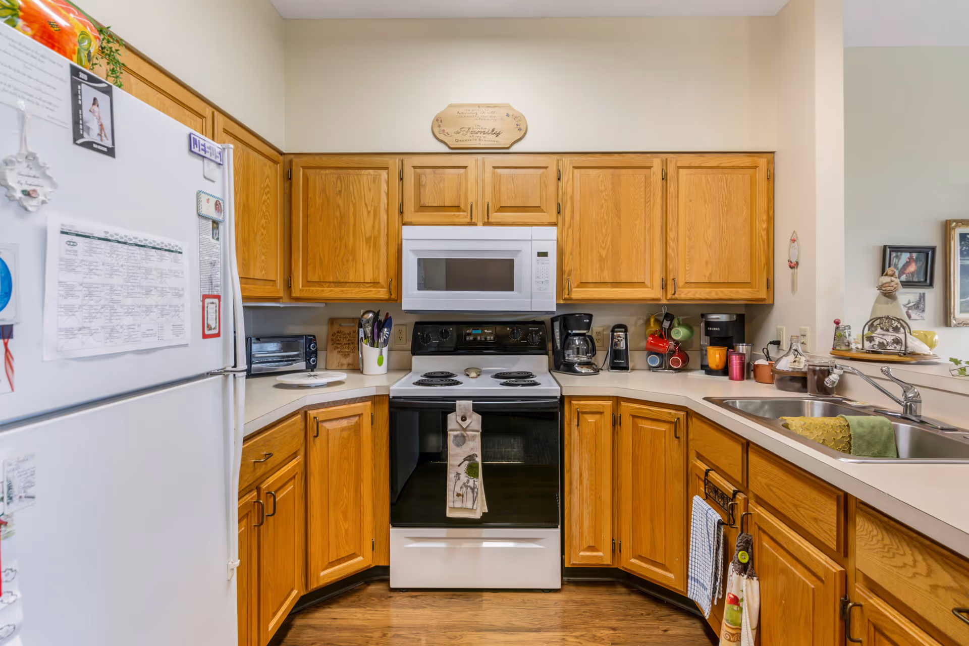 A cozy kitchen with wooden cabinets, a white refrigerator covered with magnets and papers, a white stove with a microwave above it, a coffee maker, and a double sink with a green towel. The countertops have various kitchen items including a toaster oven, utensils, and mugs. The floor is wooden and the walls are painted light beige.