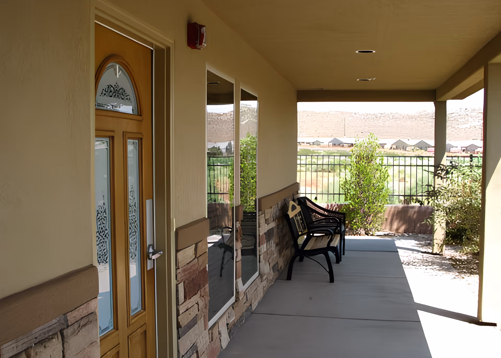 Covered outdoor patio area with a wooden door on the left, two large windows, and two black metal benches along the wall. The patio overlooks a fenced garden area with greenery and a distant view of a rocky hill.