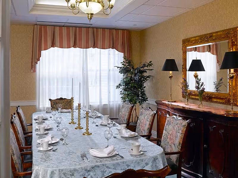 A formal dining room with a long table covered with a light blue patterned tablecloth. The table is set with white cups, plates, napkins, silverware, and glassware. There are three tall brass candlesticks with white candles in the center of the table. The room has floral upholstered chairs around the table, a large window with sheer white curtains and a striped valance, a potted plant in the corner, and a wooden sideboard with a large ornate gold-framed mirror and two black lamps with gold bases.
