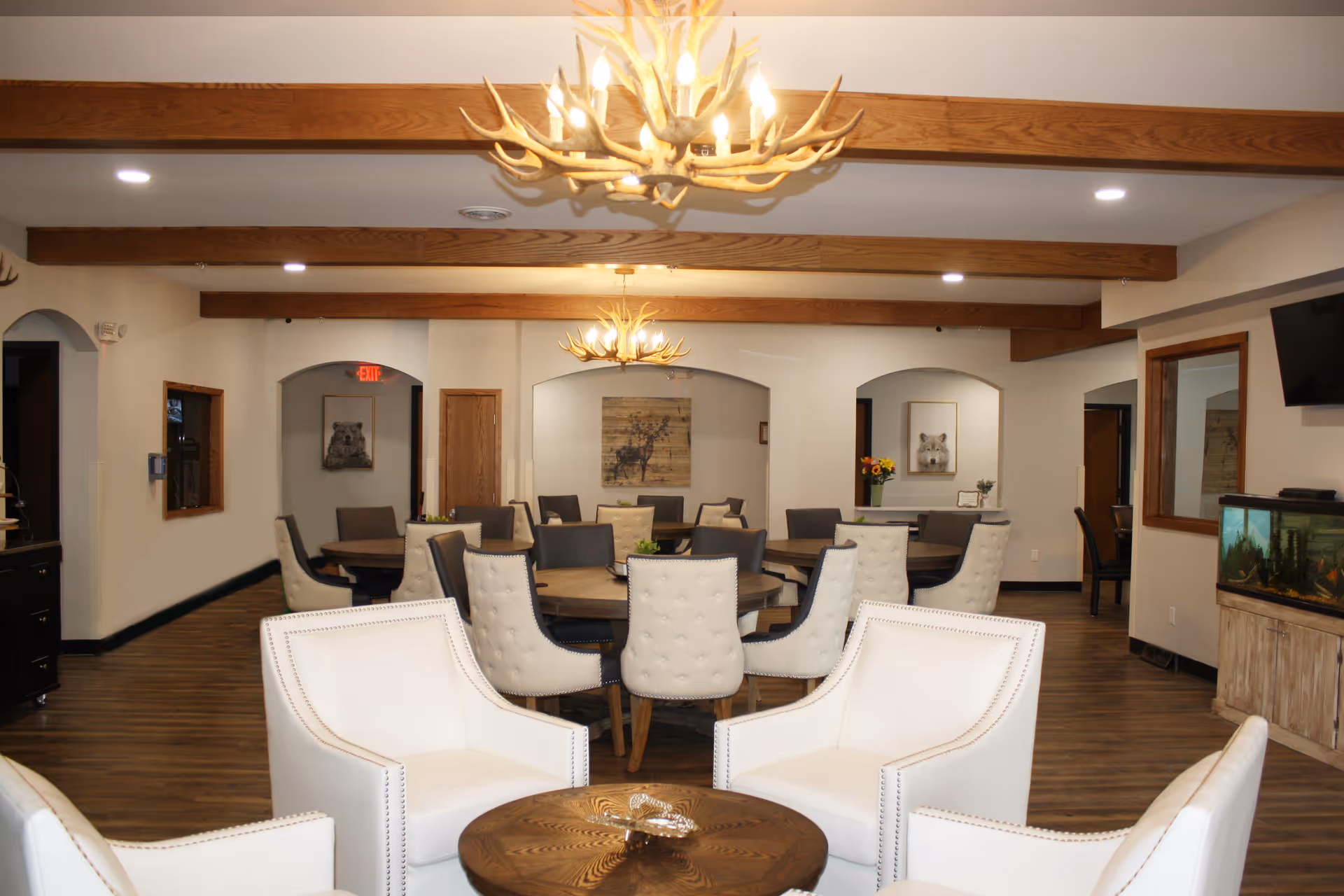 A spacious and well-lit common area in a senior living facility featuring multiple round wooden tables surrounded by cushioned chairs. In the foreground, there are four white armchairs arranged around a small round wooden coffee table. The ceiling has wooden beams and two antler-style chandeliers. The walls are decorated with framed animal artwork, and there is a fish tank on the right side of the room.