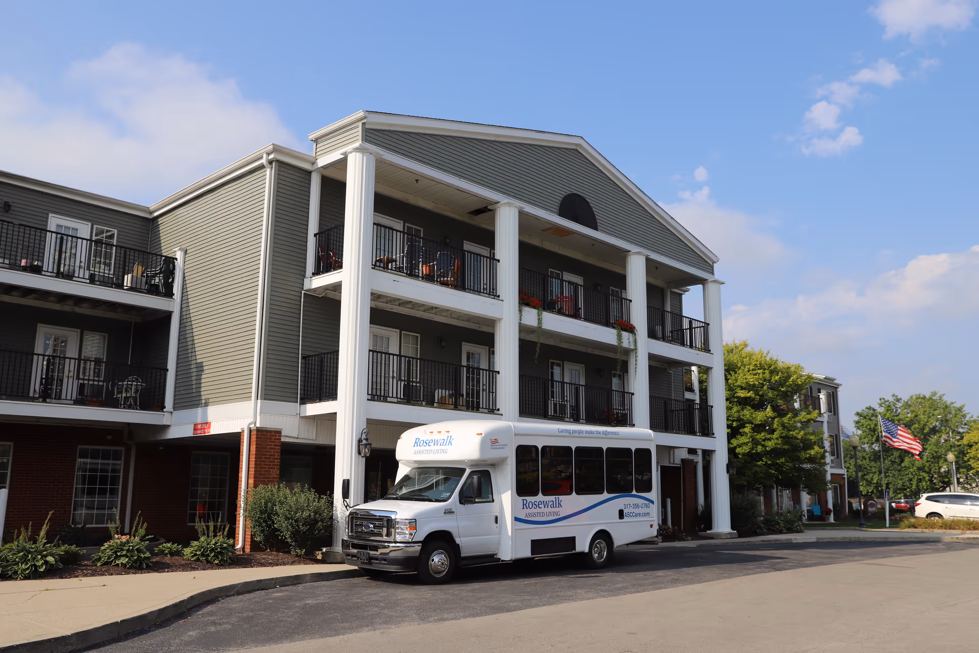 Exterior view of a three-story assisted living facility named Rosewalk at Lutherwoods, featuring balconies with black railings, white columns, and a white shuttle bus parked in front with the Rosewalk logo. The sky is partly cloudy and there is an American flag visible on the right side.