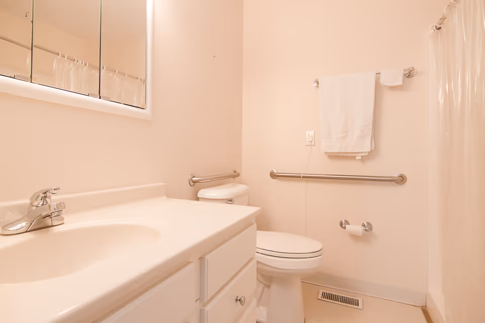 A clean and simple bathroom with a white sink and countertop, a toilet with grab bars on the wall, a towel rack with a white towel, and a shower with a clear curtain.
