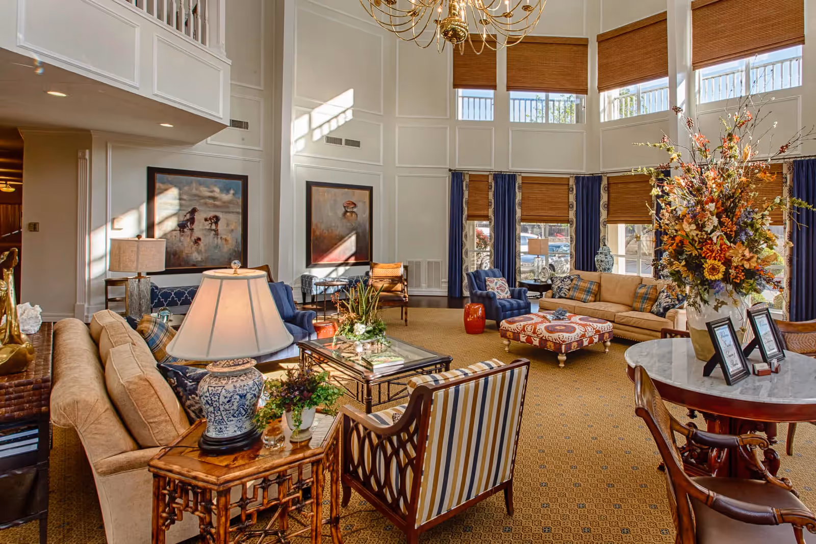 A spacious and well-lit senior living facility common area with high ceilings and large windows covered by brown blinds. The room features comfortable seating including beige sofas, blue armchairs, and a patterned ottoman. There are decorative elements such as a large floral arrangement on a round table, table lamps, framed artwork on the walls, and various plants. The carpet is a warm golden color with a subtle pattern.