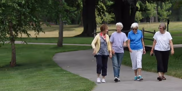 Four elderly women walking and talking together on a paved path in a park-like outdoor setting with green grass and trees.