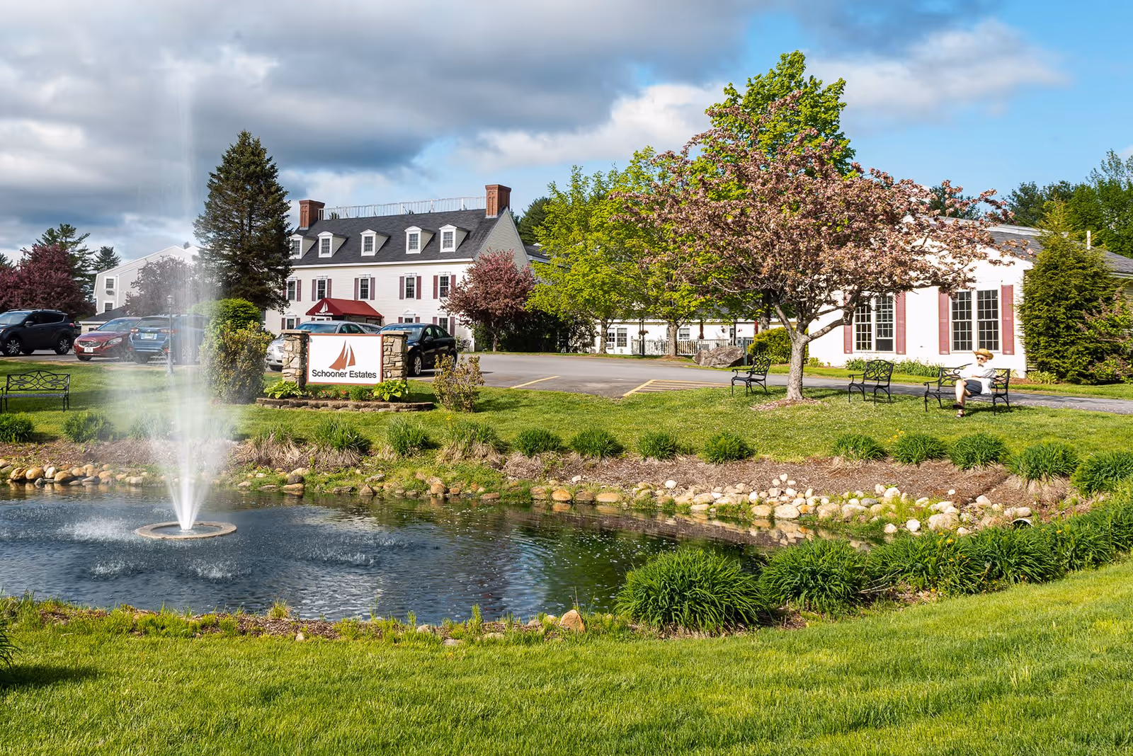 Outdoor view of Schooner Estates Senior Living featuring a small pond with a water fountain in the foreground, green grass and landscaped plants surrounding the pond, a parking area with several cars, a large white building with multiple windows and a black roof in the background, and a person sitting on a bench under a tree with pink blossoms.