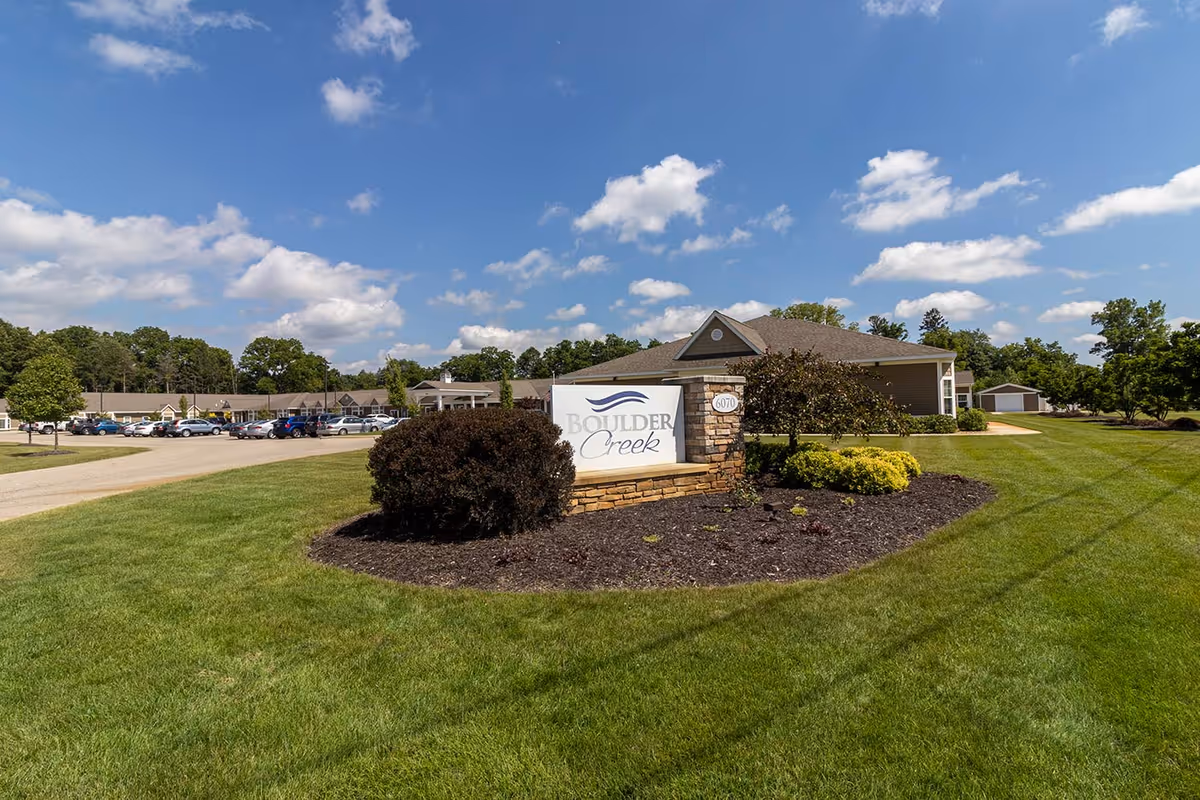 Exterior view of Boulder Creek Assisted Living & Memory Care facility with a large green lawn, a stone sign displaying the facility name, a parking lot with cars, and a partly cloudy blue sky.