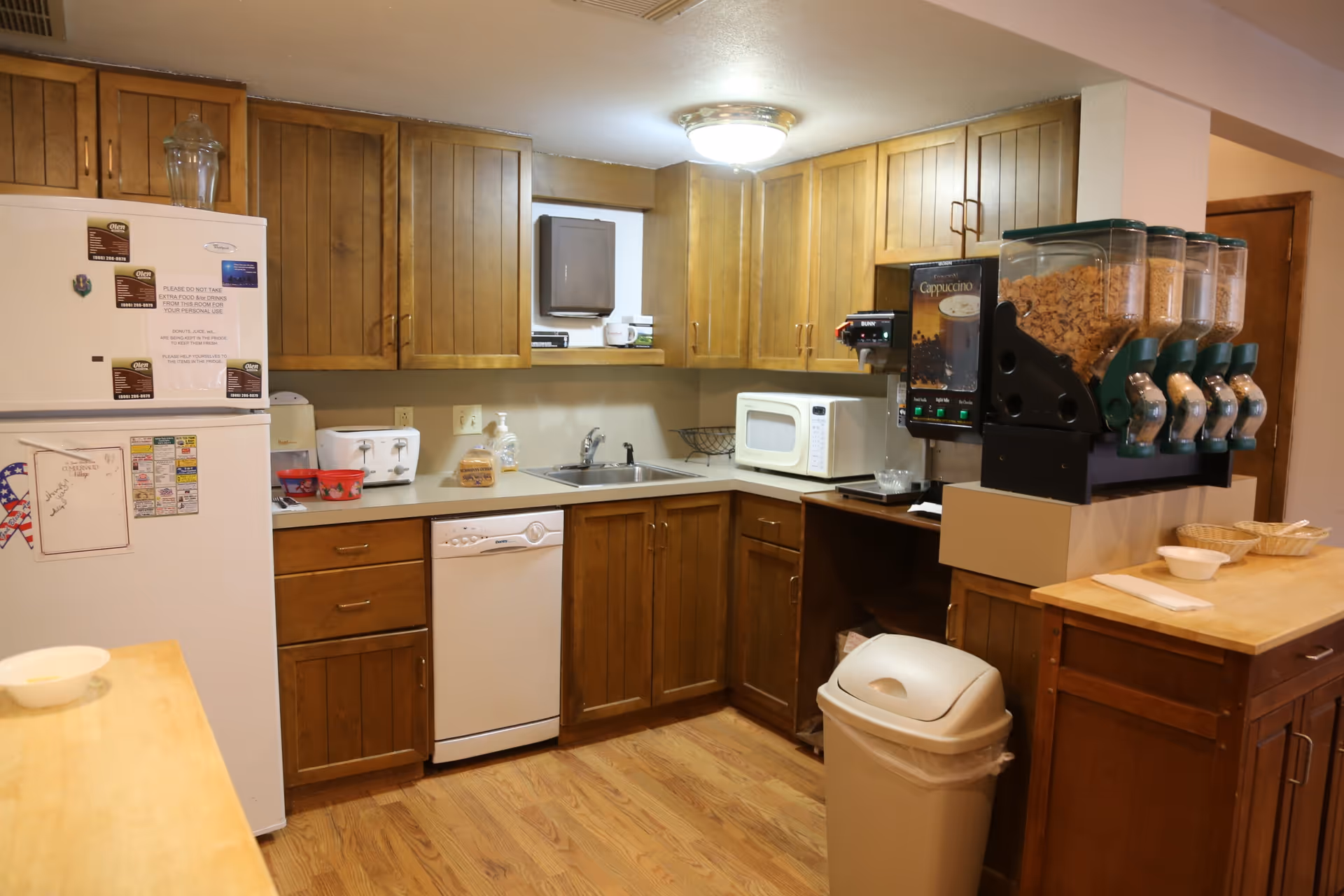 A small kitchen area with wooden cabinets, a white refrigerator with notes and magnets, a dishwasher, a sink, a microwave, a toaster, a coffee and cereal dispenser, and a trash can. The floor is wooden and the lighting is warm.